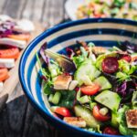 A vibrant mixed green salad featuring cherry tomatoes, cucumber, croutons, and spring onions is presented in a blue-striped bowl, accompanied by sliced tomato, mozzarella, and red onion on a wooden board.