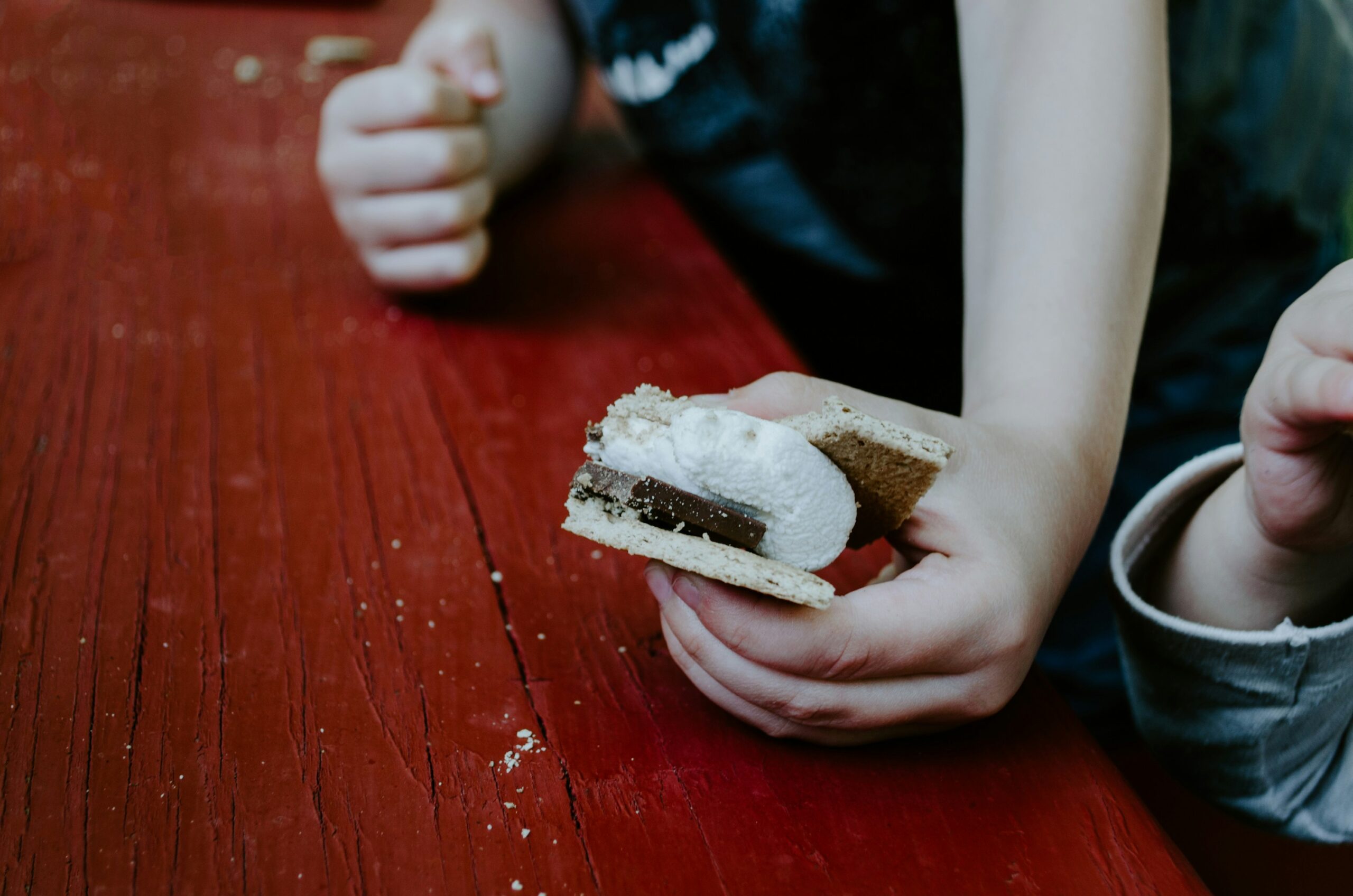 On a rustic red wooden table, a child is holding a homemade s'more prepared with chocolate and melted marshmallow between graham crackers.
