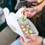 A person holds chopsticks and a sushi roll over a takeout box containing sushi rolls topped with sesame seeds and sauce.