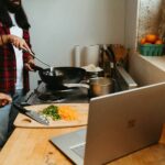 While following a recipe on a laptop, a person is cooking with a wok on a burner. In the vicinity, a cutting board contains chopped veggies.