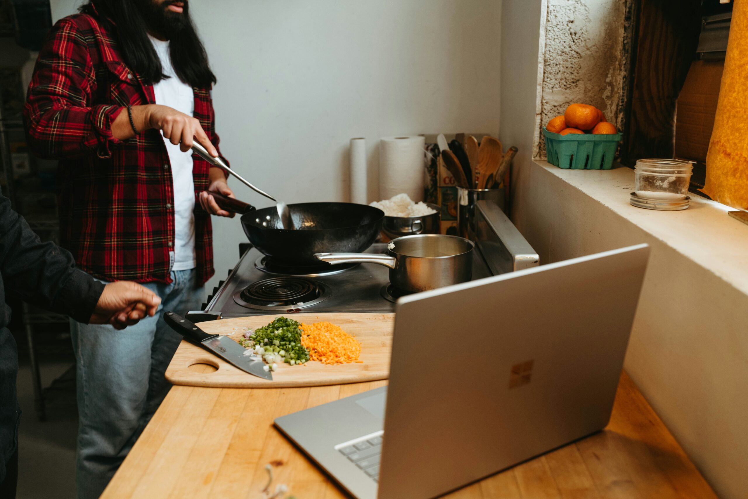 While following a recipe on a laptop, a person is cooking with a wok on a burner. In the vicinity, a cutting board contains chopped veggies.