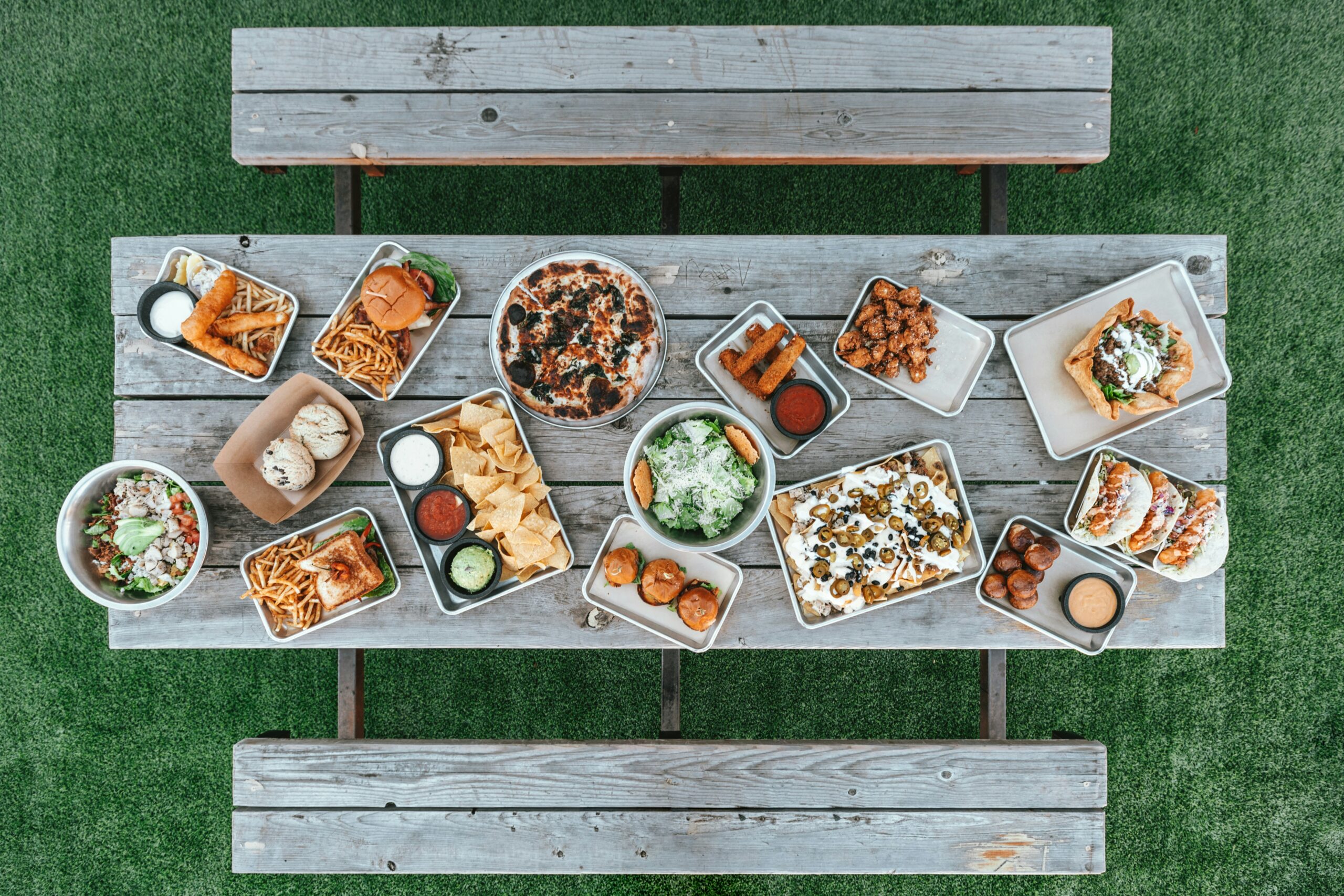 A top-down overhead view of a wooden picnic table filled with various adult happy meal-style comfort foods, including burgers, tacos, nachos, pizza, and appetizers on a green grass background.