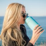 Blonde woman with sunglasses drinking from a large turquoise insulated water bottle by the ocean, profile view on a sunny beach day promoting hydration and healthy lifestyle