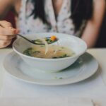 A person holding a spoon over a white bowl of clear vegetable soup containing noodles, carrots, and green onions.