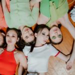 Spread out like petals, five girls rest on a wooden floor, faces pointing straight ahead. Their heads meet in the middle, bodies fanning outward in bright splashes of color - red, sharp green, crisp white. Makeup shaped with care frames quiet eyes fixed on the lens. A stillness holds them, posed without motion. The wood beneath carries no sound