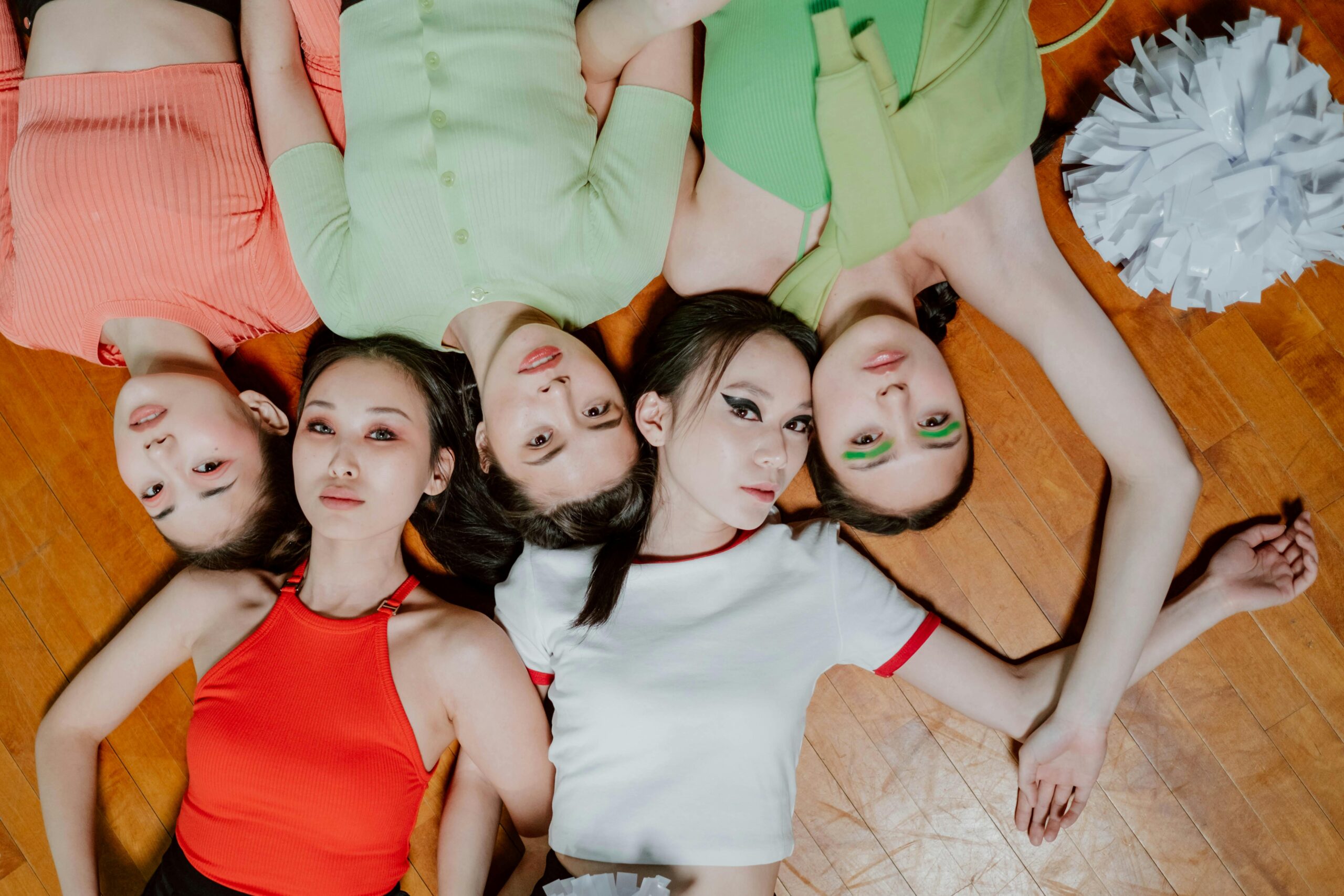 Spread out like petals, five girls rest on a wooden floor, faces pointing straight ahead. Their heads meet in the middle, bodies fanning outward in bright splashes of color - red, sharp green, crisp white. Makeup shaped with care frames quiet eyes fixed on the lens. A stillness holds them, posed without motion. The wood beneath carries no sound