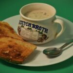 Beside it lies a flat dish, empty except for support. Two pieces of bread sit close by, edges crisped, color deepened by heat. A utensil made of metal waits nearby, polished but unused. Words curve across the cup in faded blue ink - “SCOTCH BROTH” - styled like old print. Tiny drawings circle the label: roots, greens, things pulled from soil. Lists follow, small and precise, naming what went inside. All of it stands on a field of bright green, unbroken, bold behind the scene.