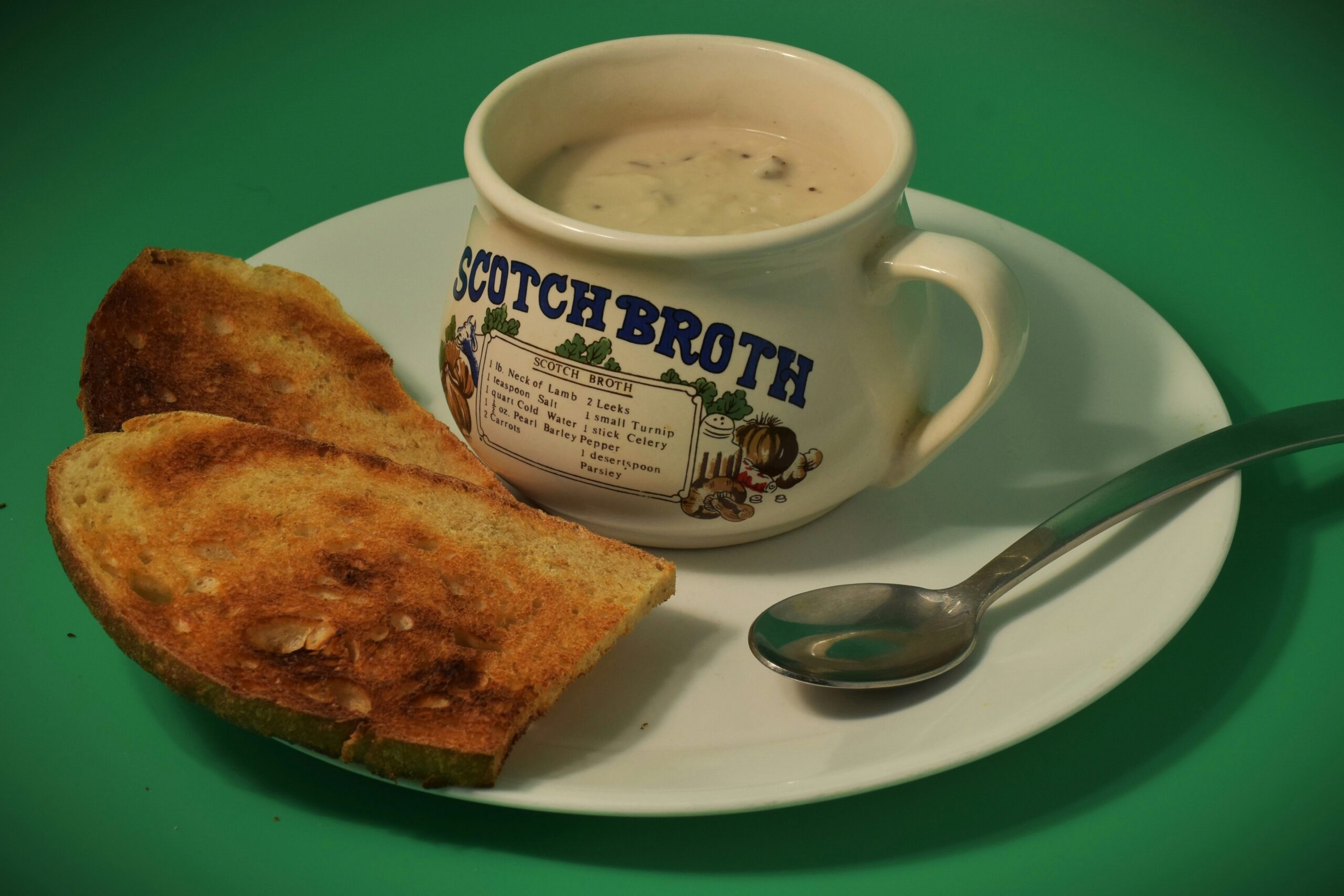 Beside it lies a flat dish, empty except for support. Two pieces of bread sit close by, edges crisped, color deepened by heat. A utensil made of metal waits nearby, polished but unused. Words curve across the cup in faded blue ink - “SCOTCH BROTH” - styled like old print. Tiny drawings circle the label: roots, greens, things pulled from soil. Lists follow, small and precise, naming what went inside. All of it stands on a field of bright green, unbroken, bold behind the scene.