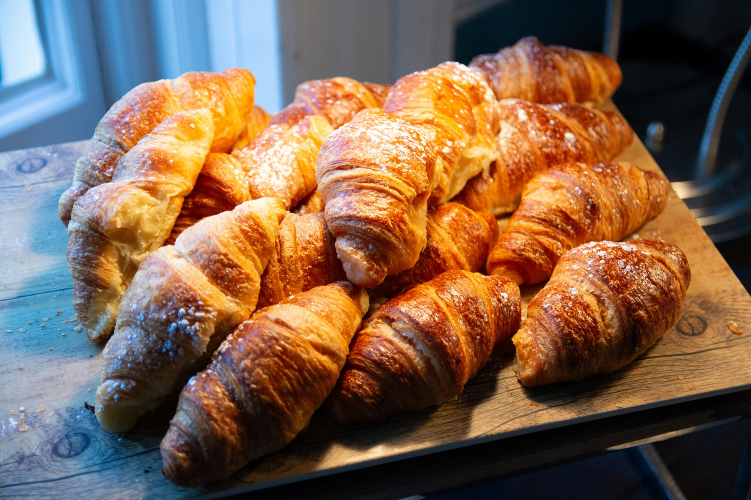 A heap of golden-brown, flaky croissants dusted with powdered sugar, resting on a wooden board with soft lighting from a nearby window.