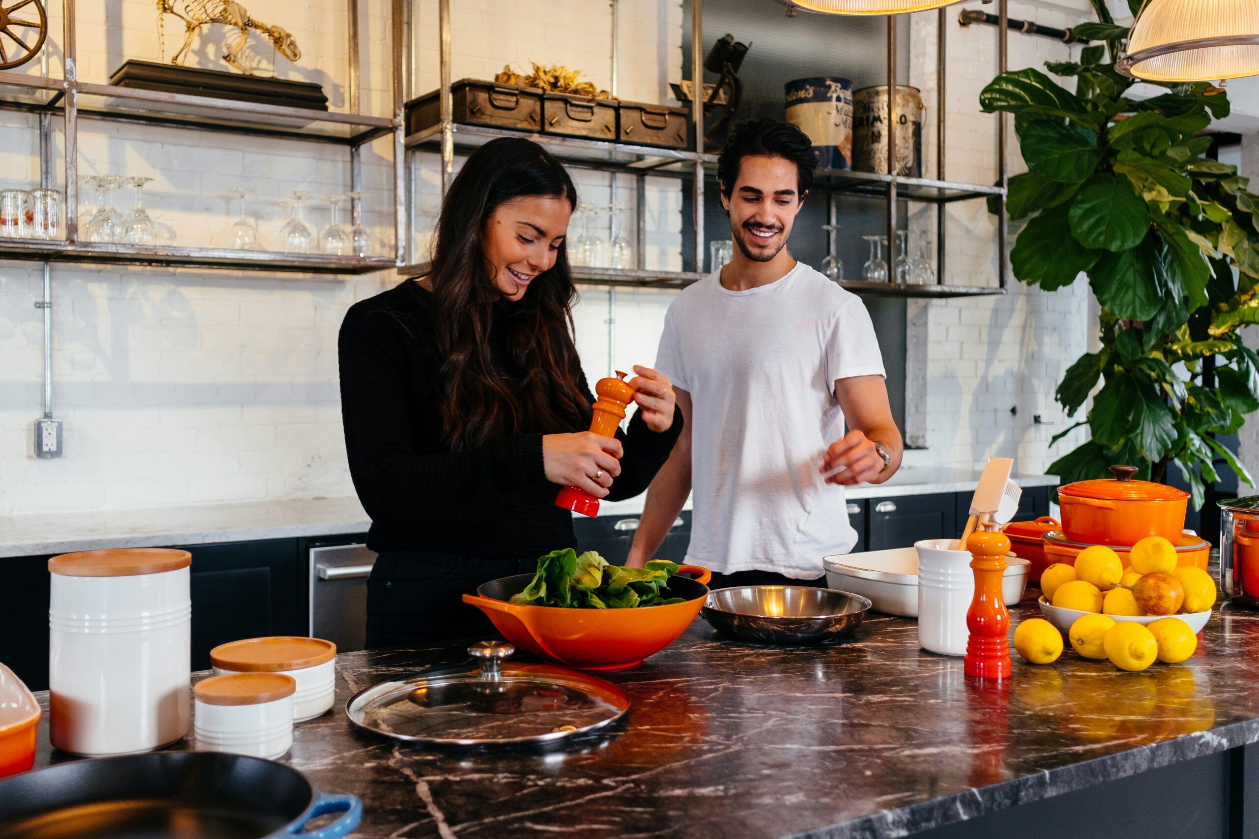 A young man and woman smiling while cooking together in a modern kitchen with industrial shelving and marble countertops.